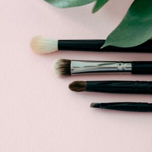 Aesthetic flat lay of makeup brushes with green leaves on a soft pink background.