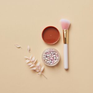 Elegant flat lay of a makeup brush, container with powder balls, and dried leaves on yellow pastel surface.