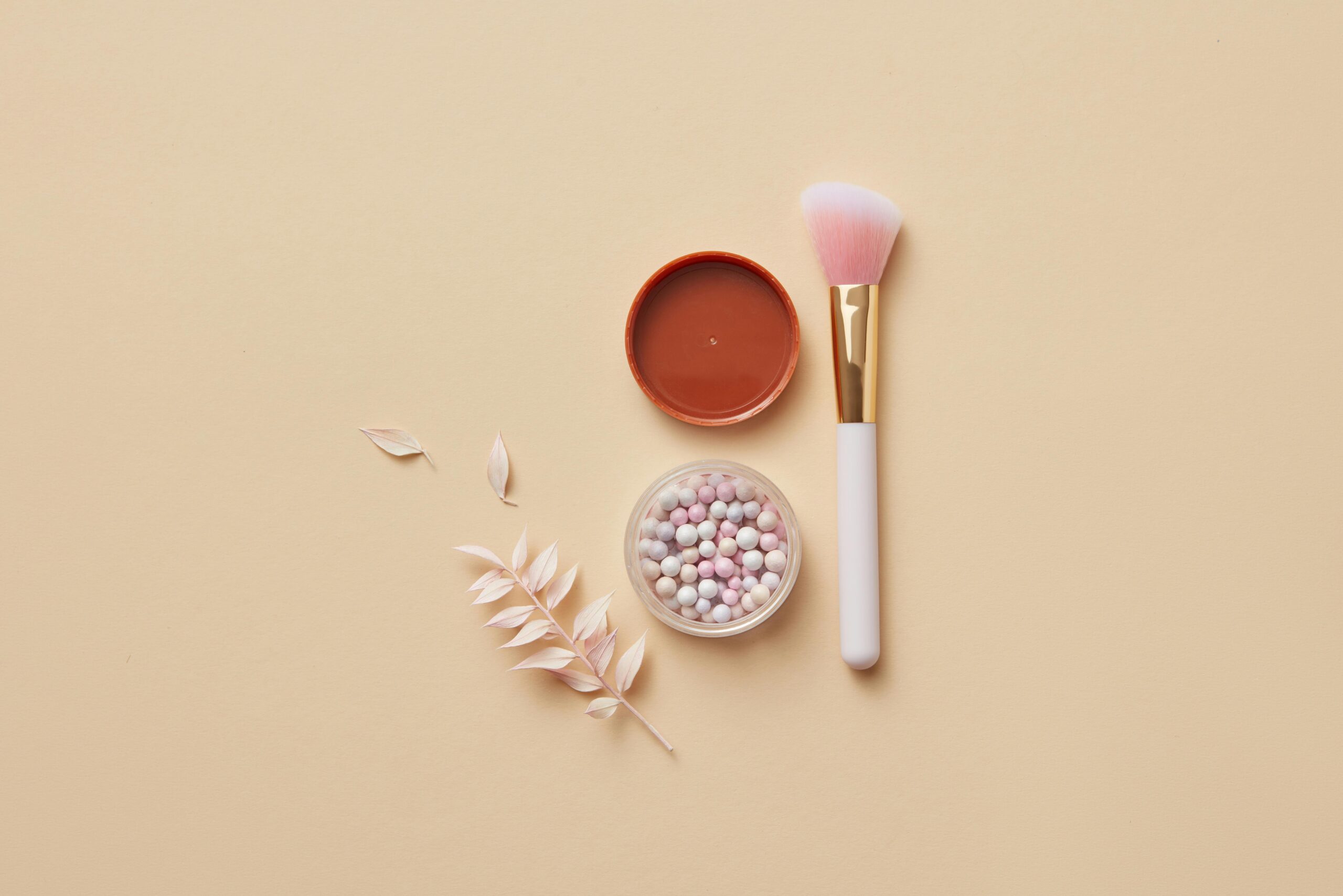 Elegant flat lay of a makeup brush, container with powder balls, and dried leaves on yellow pastel surface.
